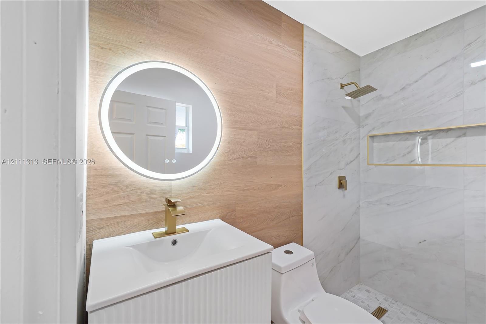 Bathroom featuring a vanity with wood-finish wall paneling, a backlit circular mirror, a gold-toned single-handle faucet, stone-surround shower with a built-in recessed niche, and gold-toned rainfall shower head