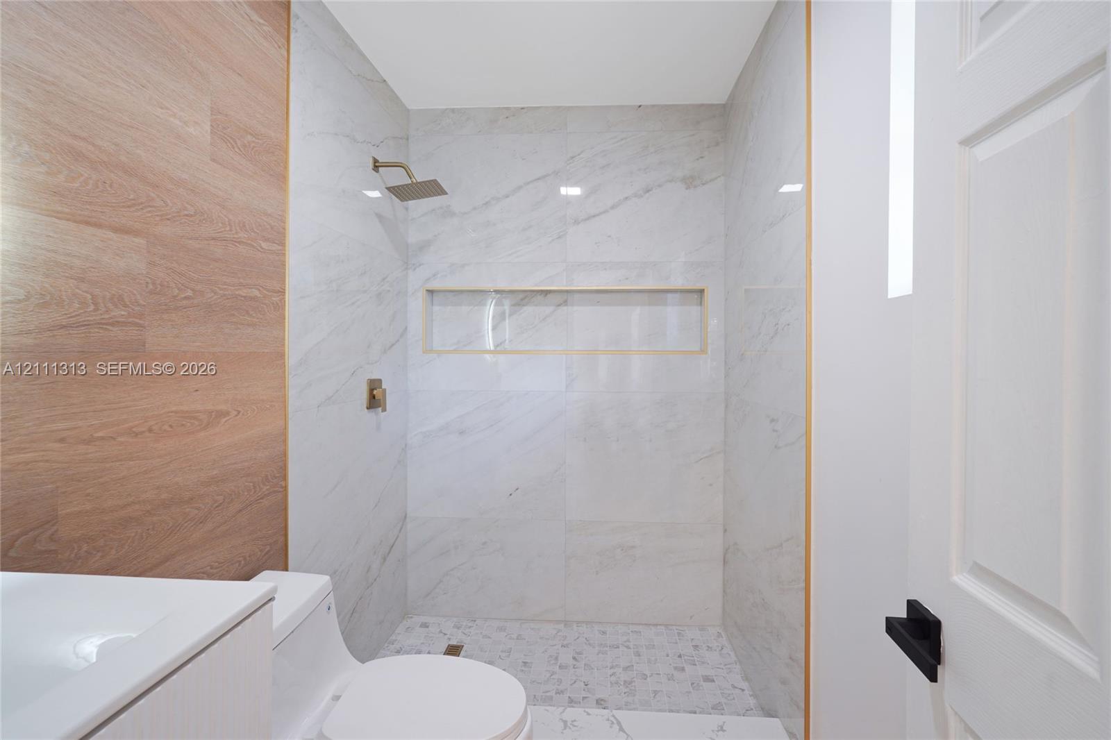Walk-in shower featuring large-format light-veined tile, wood-finish accent wall, built-in recessed shelf, rainfall shower head, and decorative metallic trim