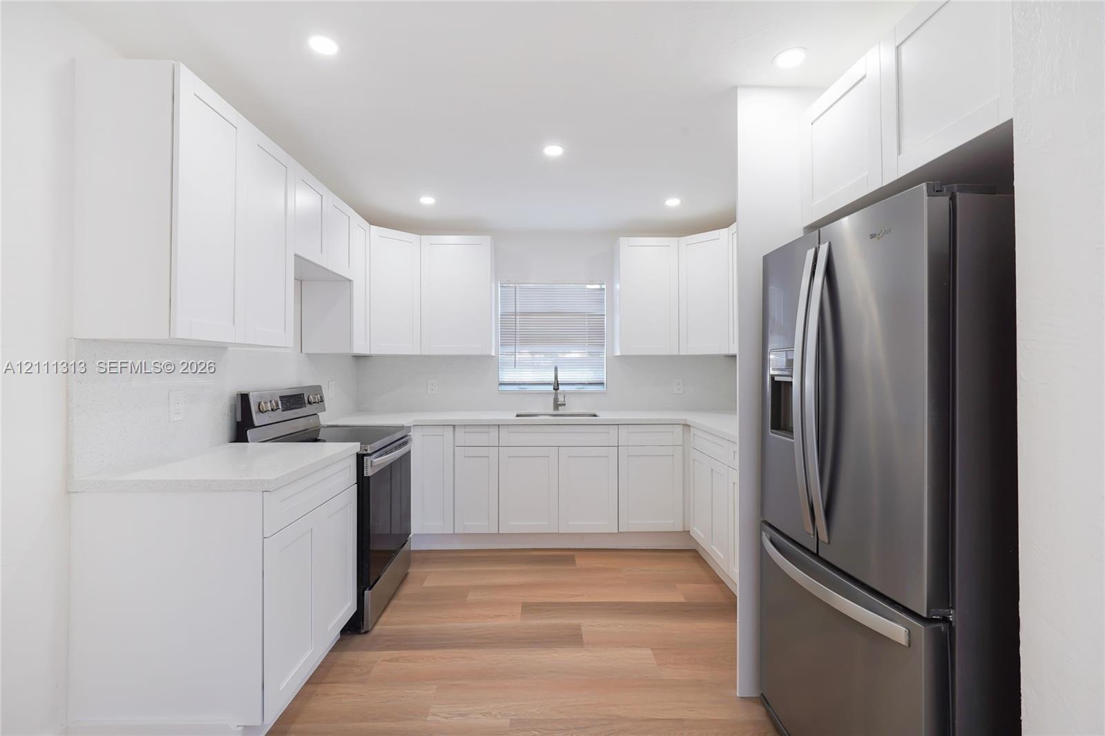 White Shaker-style cabinetry, stainless steel appliances, light-toned countertops, wood-finish flooring, and recessed lighting