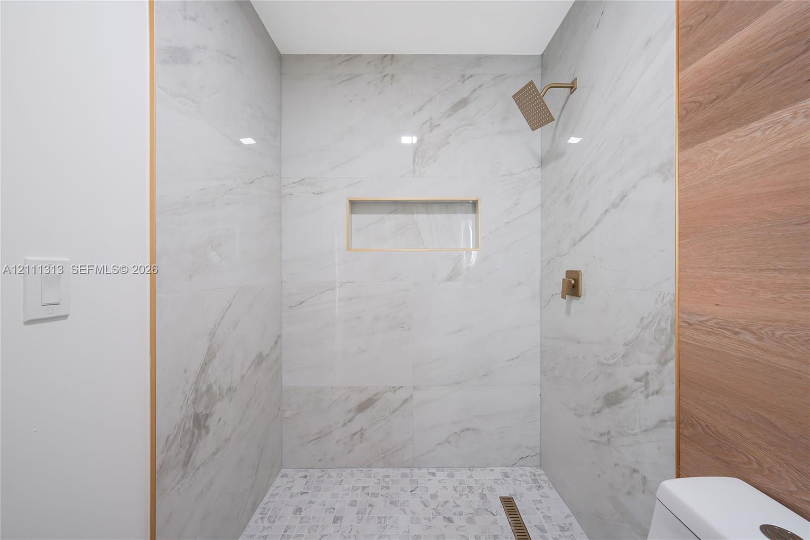 Walk-in shower featuring large-format stone-pattern tile walls, wood-finish paneling, an integrated recessed wall niche, a square rainfall showerhead, and mosaic tile flooring