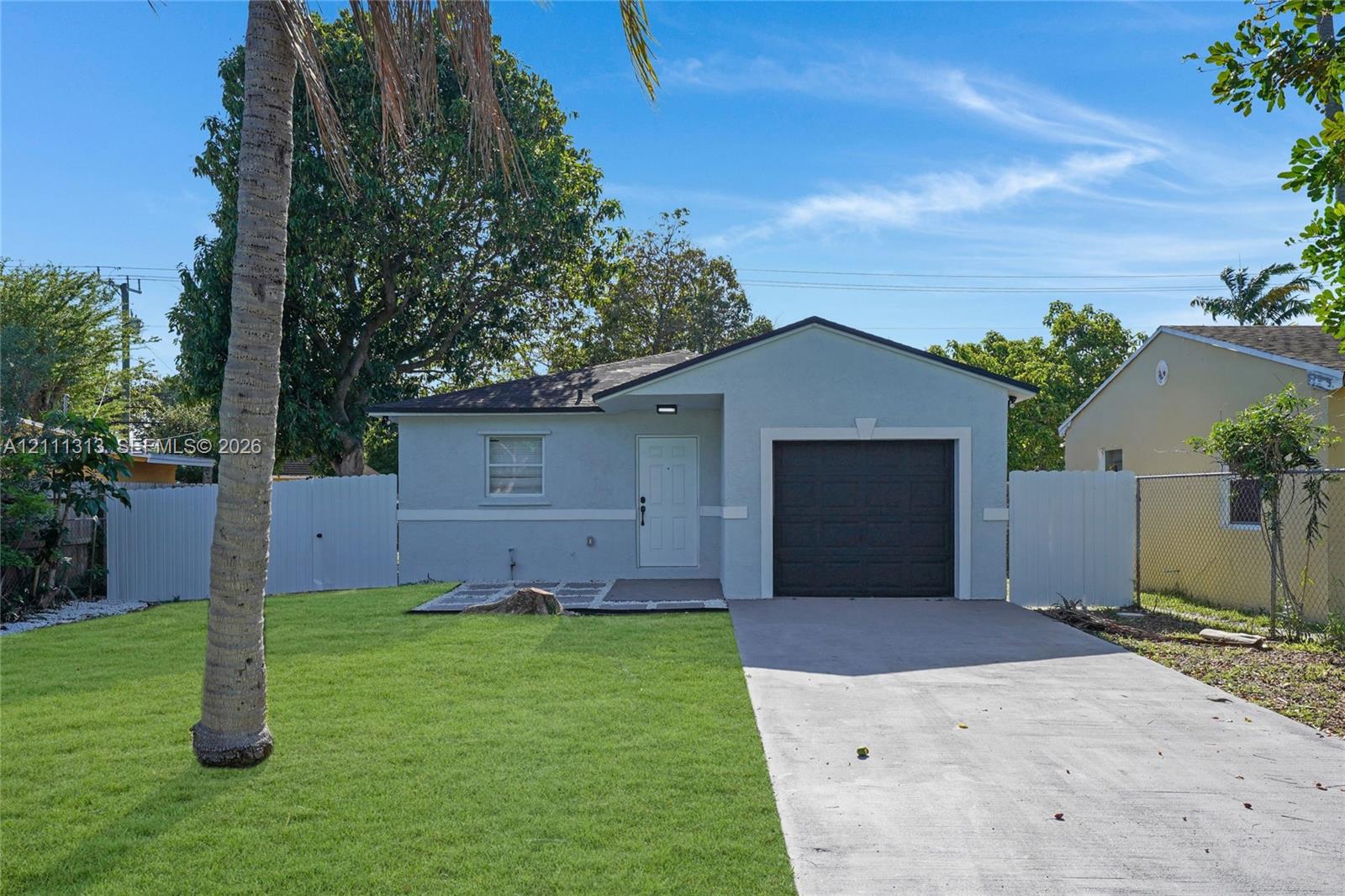 Single-story residence featuring a light-toned stucco exterior, a concrete driveway, an attached garage with a dark-finish door, and perimeter fencing
