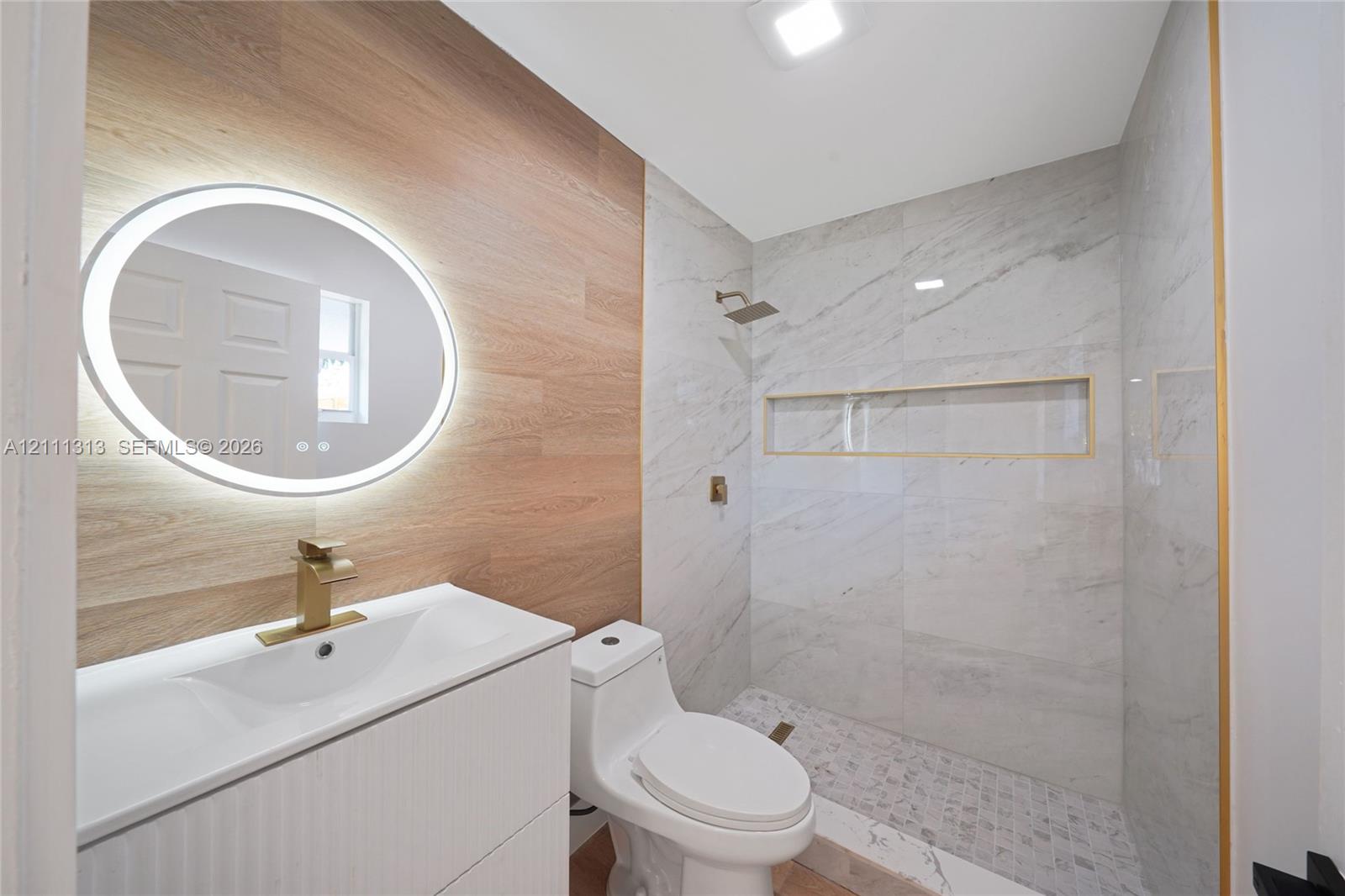 Bathroom featuring a floating vanity with a vessel sink, illuminated oval mirror, gold-tone hardware, wood-finish wall paneling, and a tiled shower with a built-in recessed niche