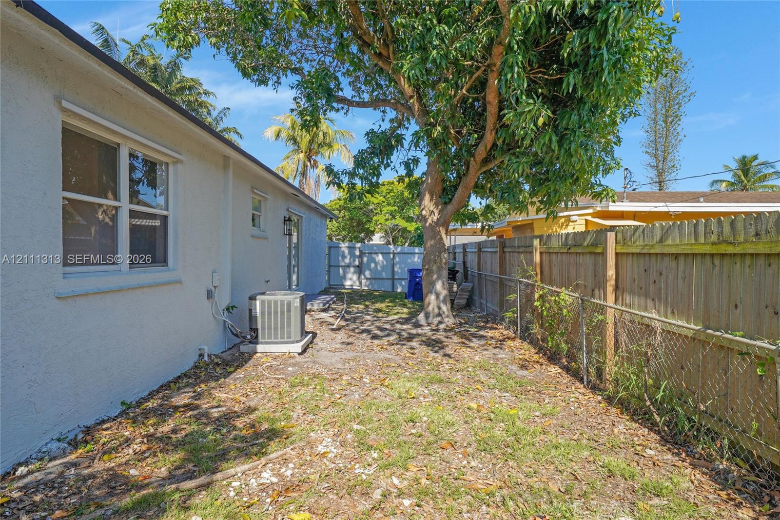 Exterior side yard featuring stucco siding, double-hung windows, a mature tree, and wood-panel perimeter fencing