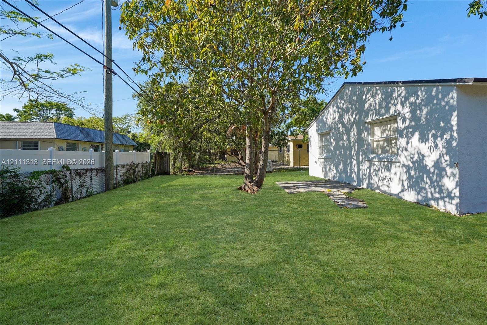 Backyard featuring a manicured lawn, stone patio, mature trees, and vinyl perimeter fencing