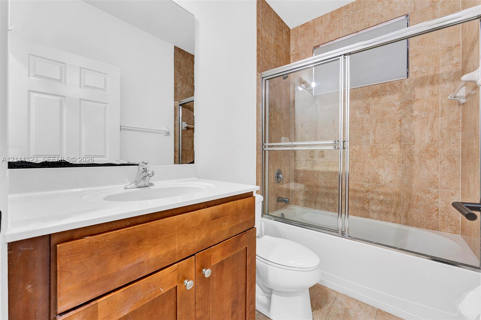 Wood-finish vanity with a solid-surface countertop, integrated sink, and chrome-finish faucet, paired with a full-width mirror and a bathtub featuring sliding glass doors and tiled surround