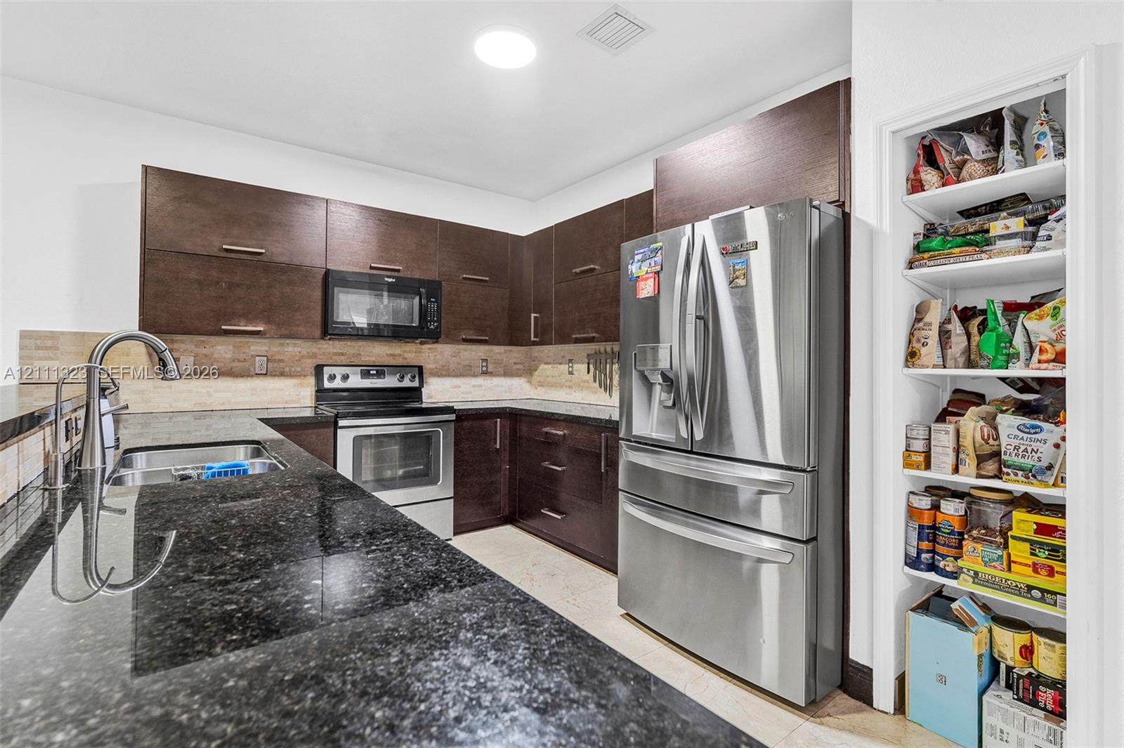 Kitchen featuring dark wood-finish cabinetry, stone-finish countertops, stainless steel appliances, a tiled backsplash, and an integrated pantry