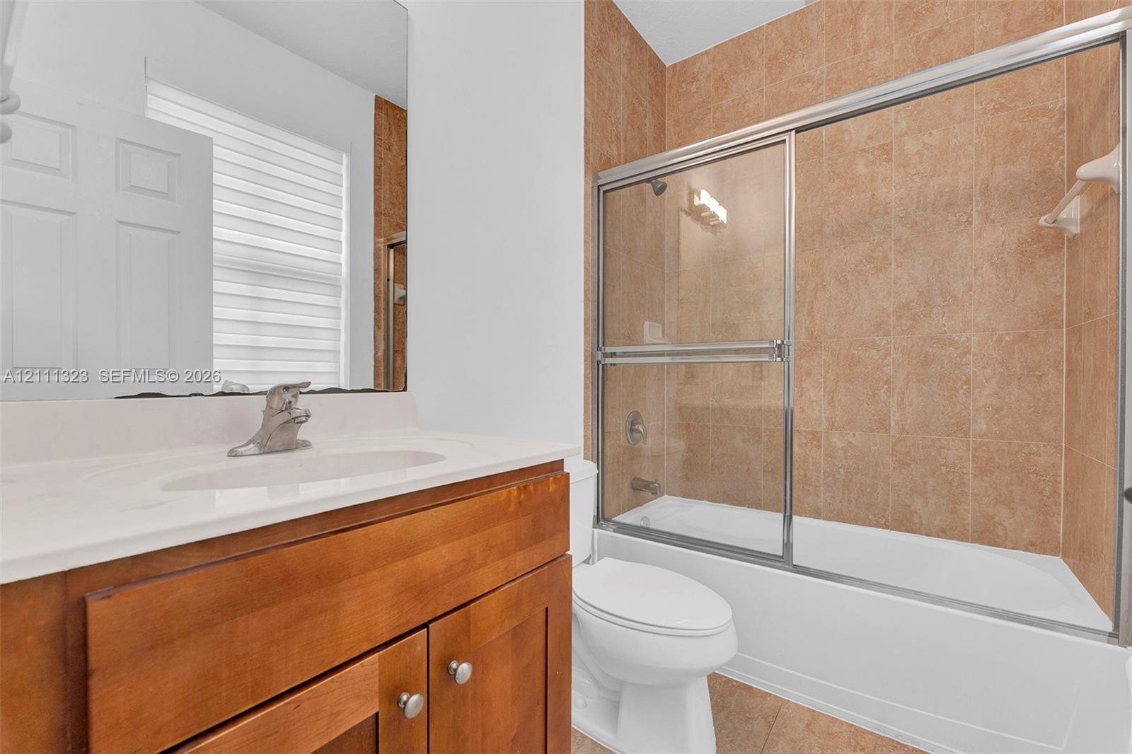 Bathroom featuring a wood-finish vanity with a white integrated sink, chrome faucet, wall-mounted mirror, and a tub-shower combination with floor-to-ceiling tile surround and sliding glass doors