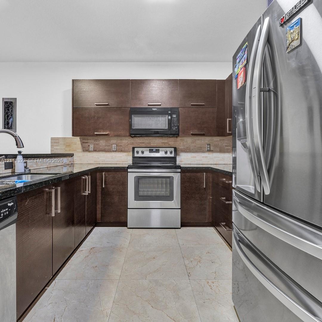 Kitchen featuring dark wood-finish cabinetry, stainless steel appliances, dark stone countertops, tile backsplash, and light-toned tile flooring