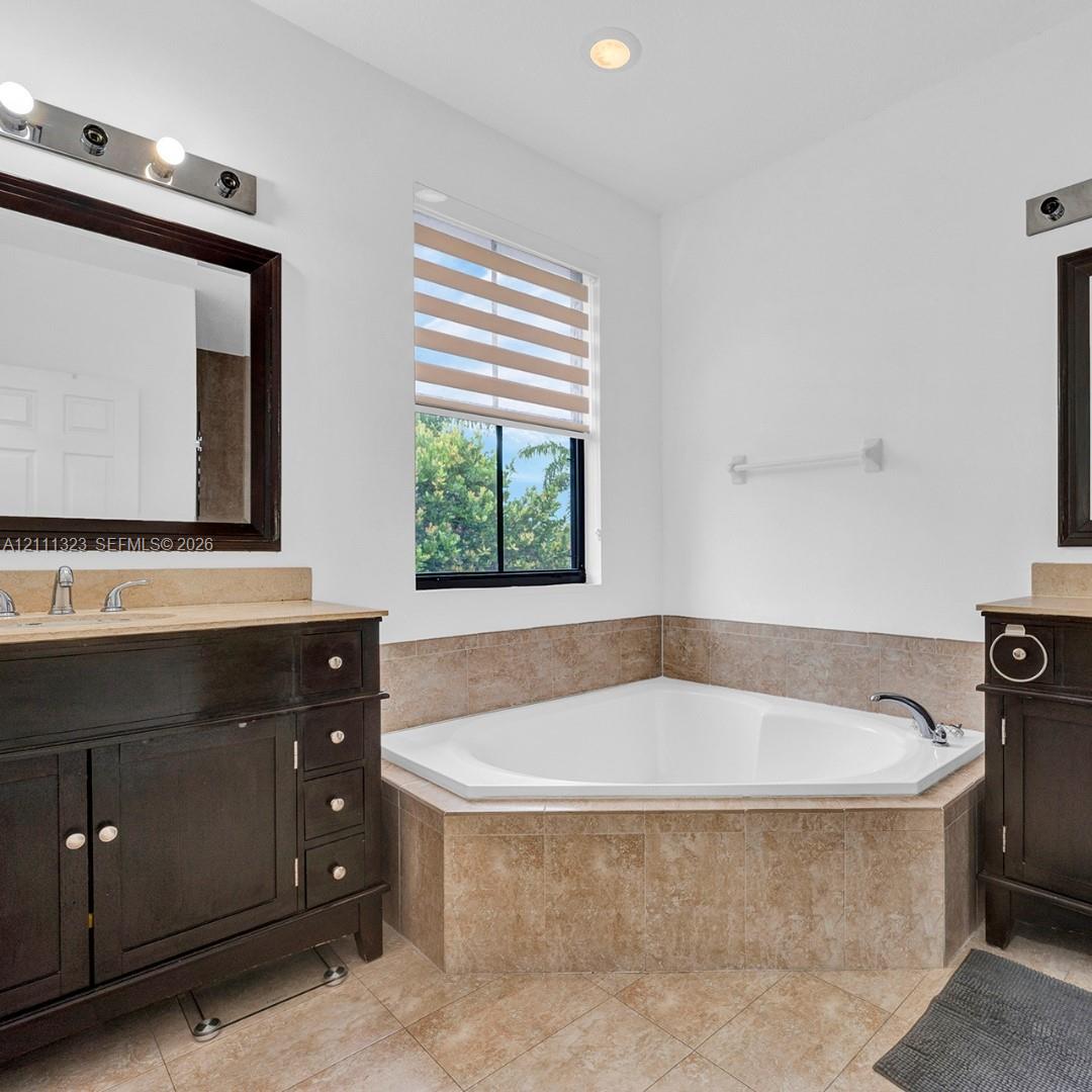 Corner soaking tub with tile surround, dual dark-finish wood cabinetry, natural stone countertops, under-mount sinks, and framed vanity mirrors