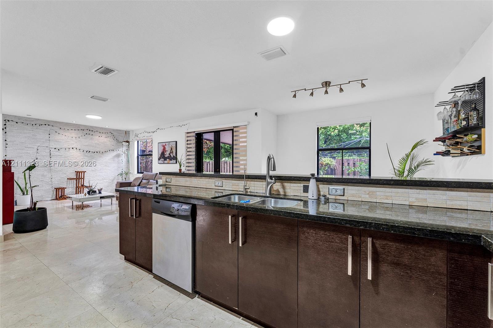 Kitchen featuring dark-toned flat-panel cabinetry, stone-finish countertops, stainless steel dual-basin sink with arched faucet, stainless steel dishwasher, and neutral tiled flooring