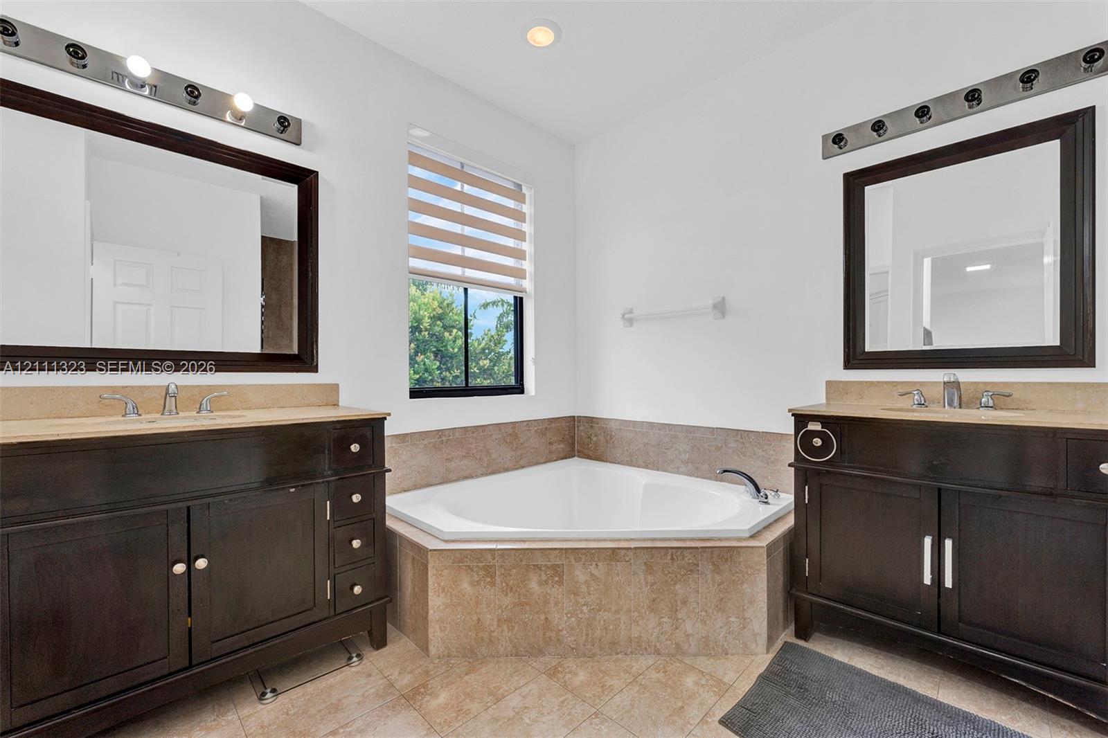 Dual dark-finish vanities with light-toned stone-style countertops, corner soaking tub with tile surround, and framed vanity mirrors