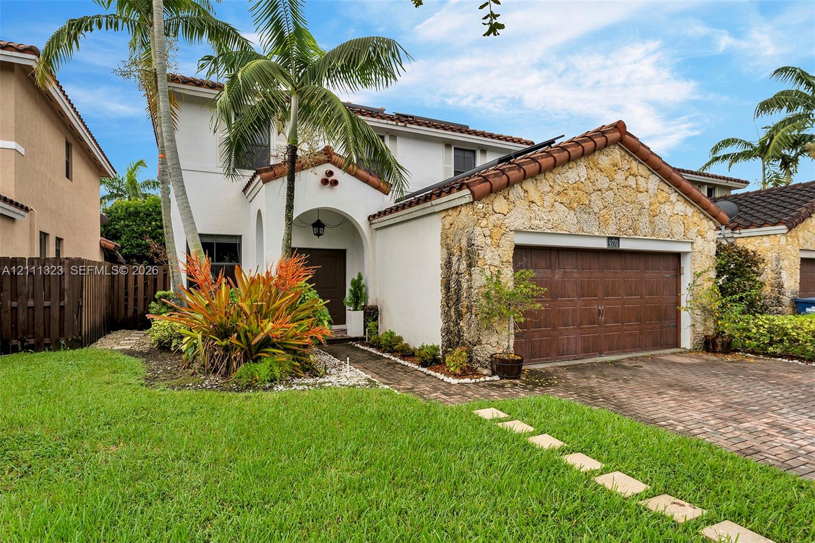 Two-story residence featuring a textured stone facade, terracotta-style roof tiles, arched entryway, and a multi-car brick-paved driveway