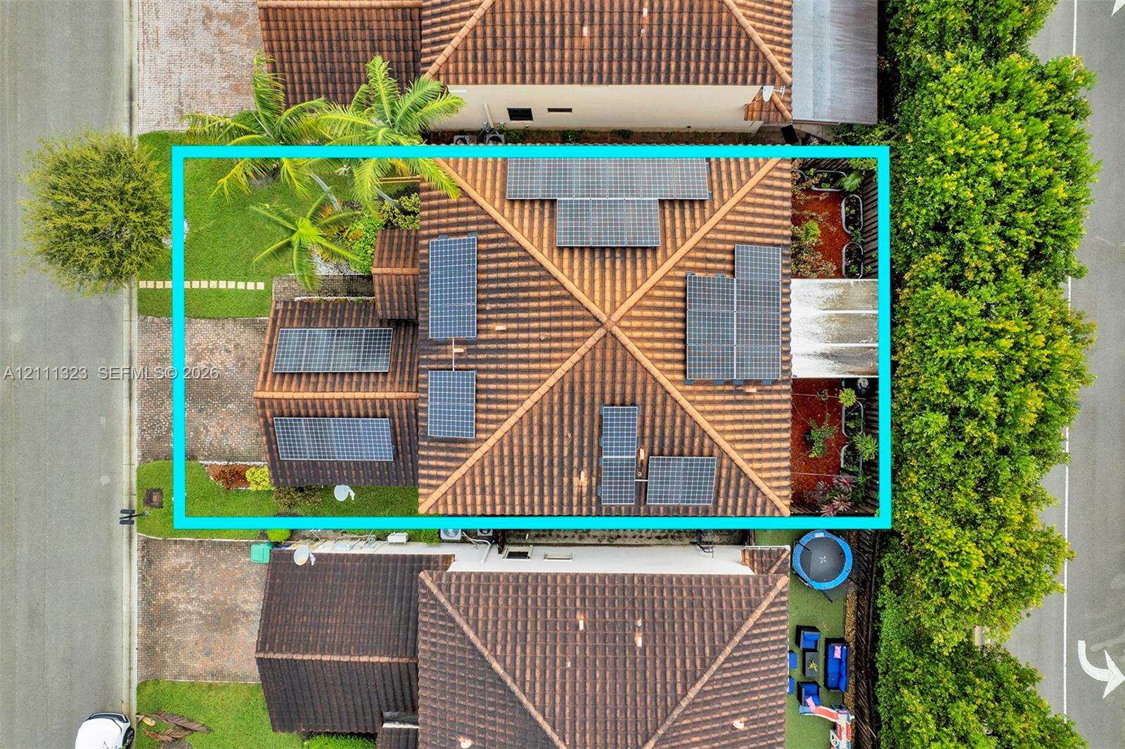Aerial view of property featuring a tile roof equipped with a solar panel array, manicured landscaping, and mature palm trees