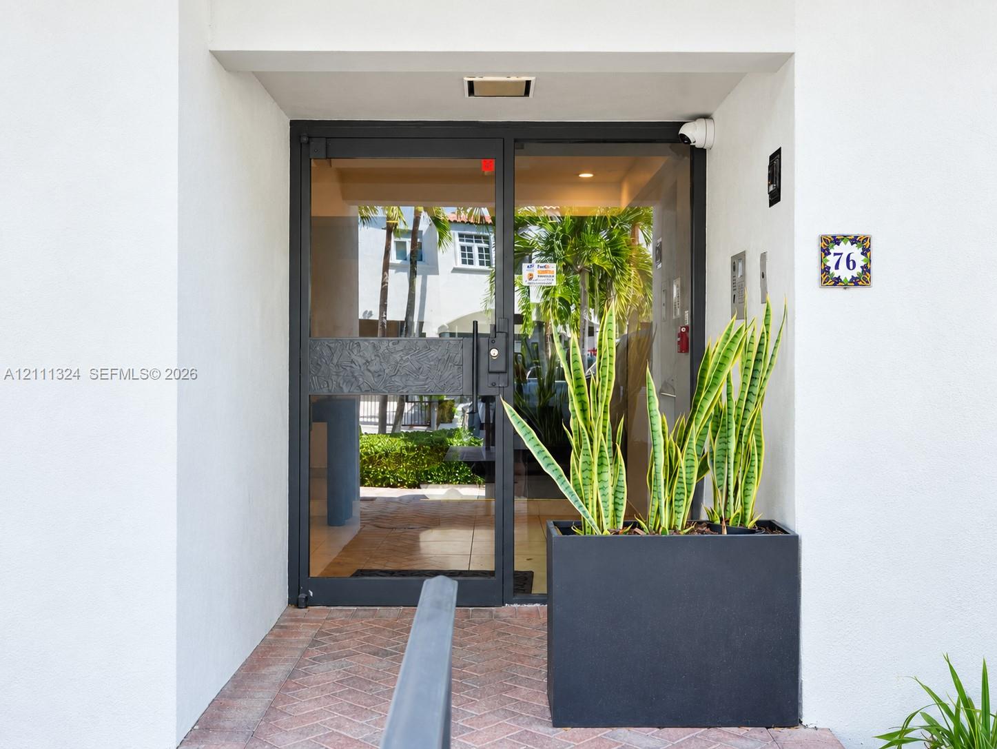 Entrance featuring a black metal-framed glass door, brick-paved walkway, recessed ceiling lighting, and a square address tile