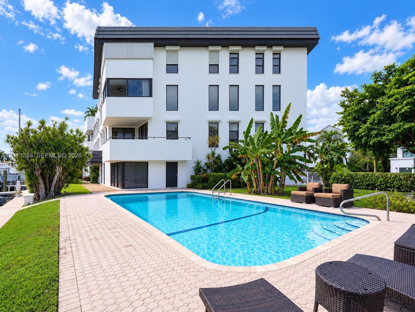 Multi-story building with a white stucco facade and charcoal-toned roofing, featuring a rectangular inground swimming pool with built-in stairs, a paved deck, and manicured landscaping