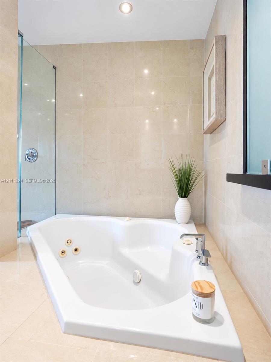 Jetted soaking tub with deck-mounted chrome faucet, recessed ceiling lighting, glass-enclosed shower, and floor-to-ceiling tile surround