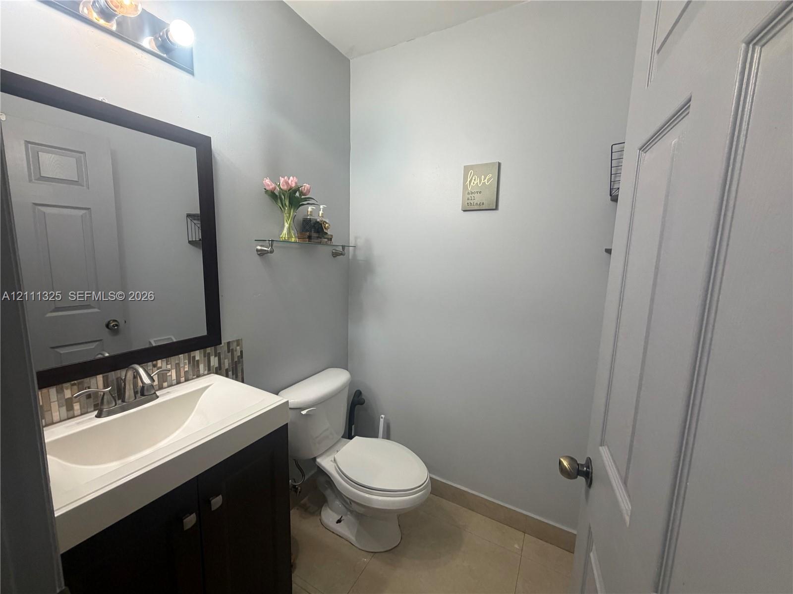 Dark wood-finish vanity with an integrated basin, glass-tile backsplash, three-light vanity fixture, and neutral tile flooring