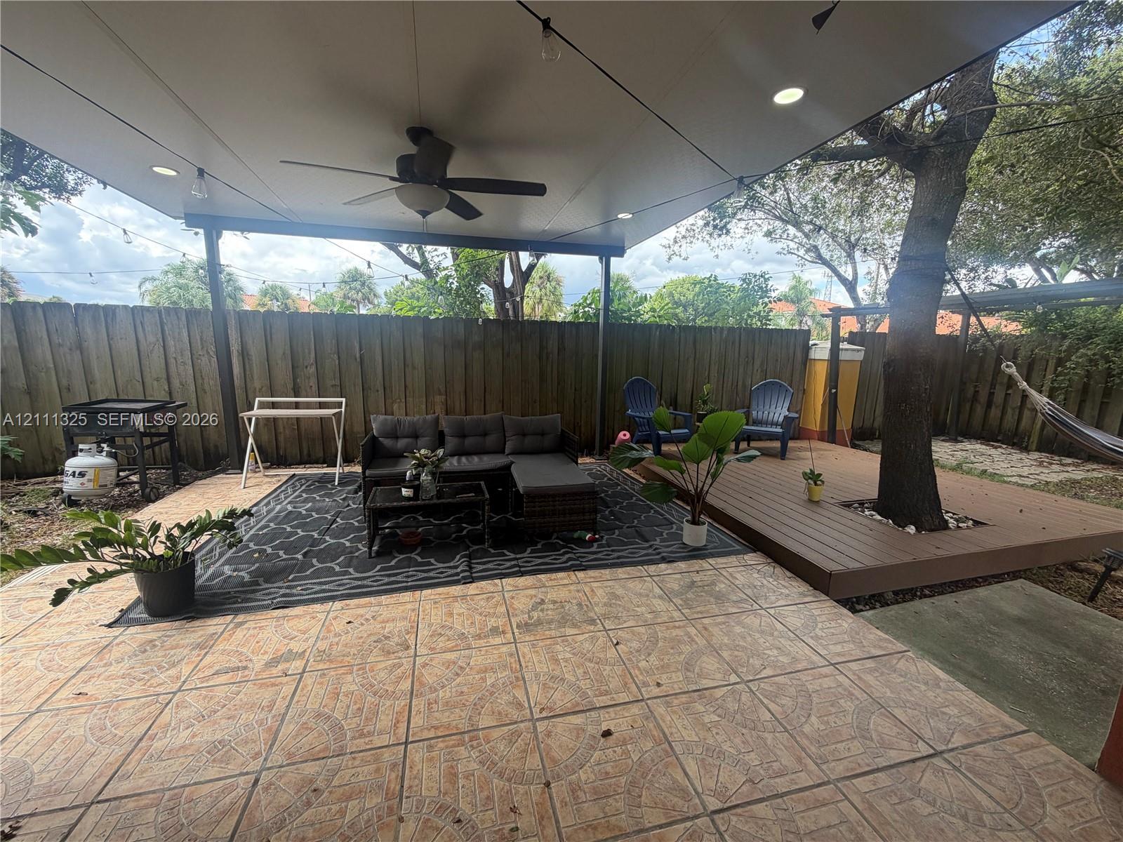 Covered patio featuring patterned tile flooring, recessed lighting, and a ceiling fan, complemented by an elevated wood-finish deck surrounding a mature tree