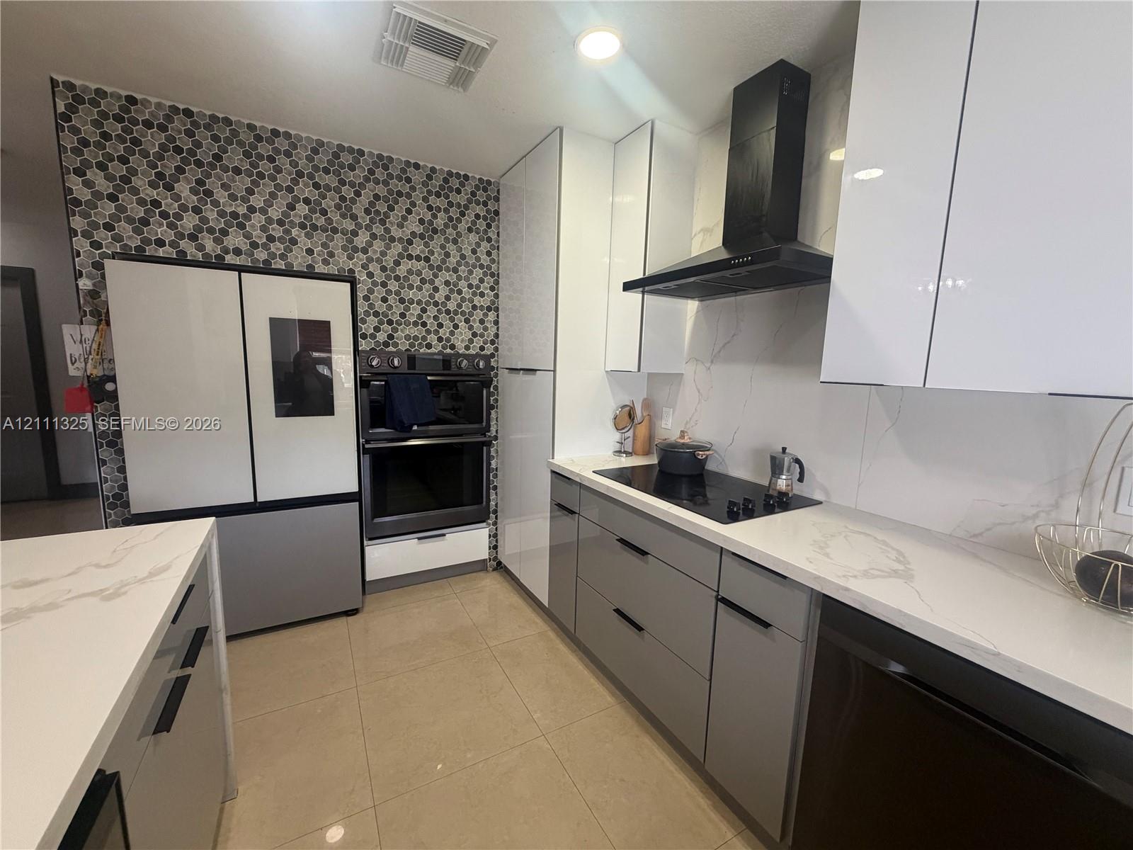 Modern kitchen featuring high-gloss cabinetry, stone-finish countertops with a full-height backsplash, hexagonal mosaic wall tiling, and a built-in double oven with a matching overhead vent hood