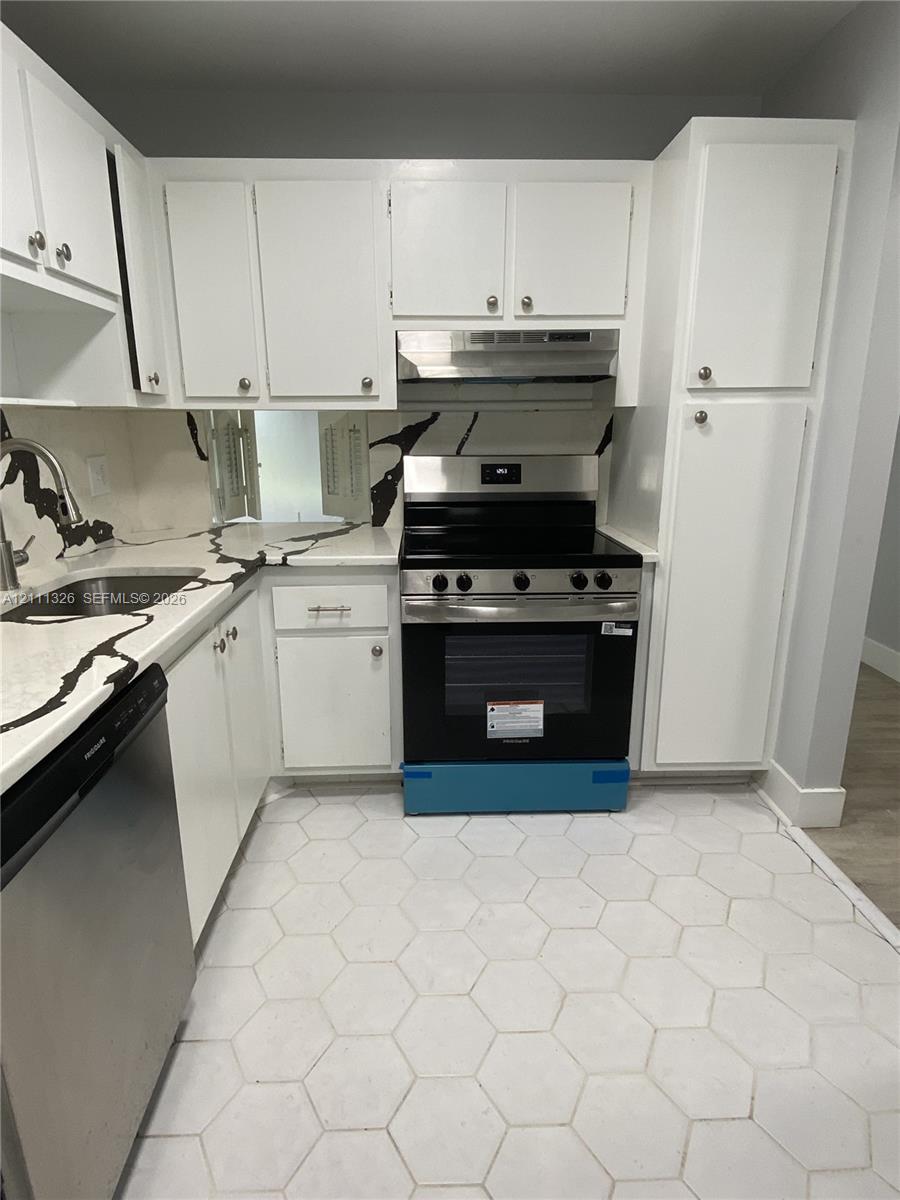 Kitchen featuring white shaker-style cabinetry, stainless steel appliances, patterned stone-finish countertops with matching backsplash, hexagonal tile flooring and passthrough to dining area.
