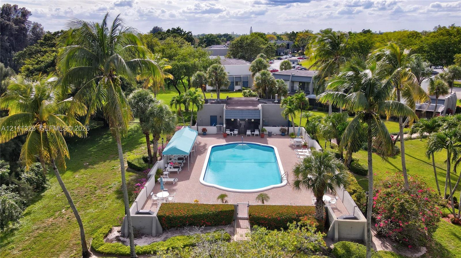 Outdoor community swimming pool with surrounding paver deck, covered patio area, and perimeter wall enclosure situated within a landscaped setting of mature trees.