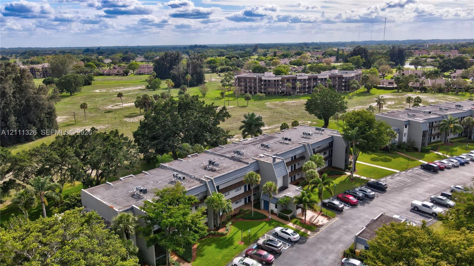 Aerial perspective featuring multi-story residential buildings, paved surface parking, manicured landscaping, and mature trees.