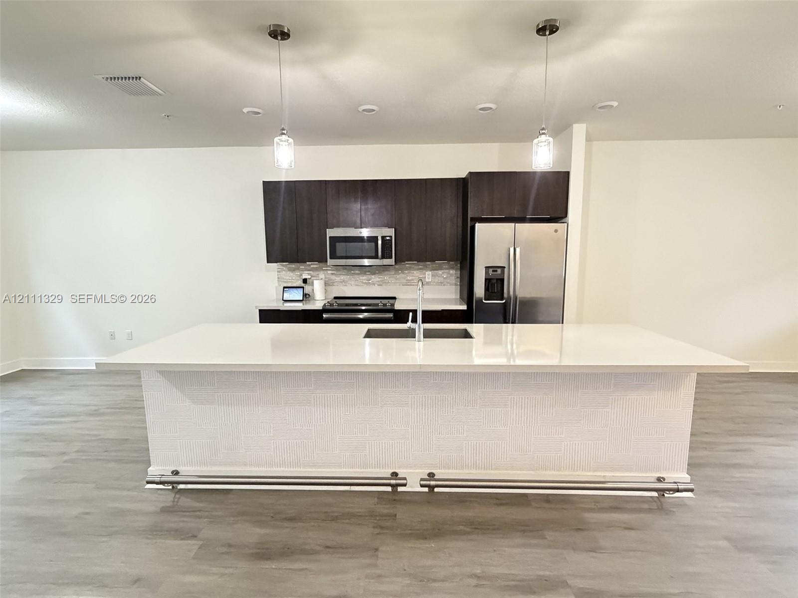 Kitchen featuring an expansive island with a solid-surface countertop, dark cabinetry, stainless steel appliances, textured island base finish, and pendant lighting