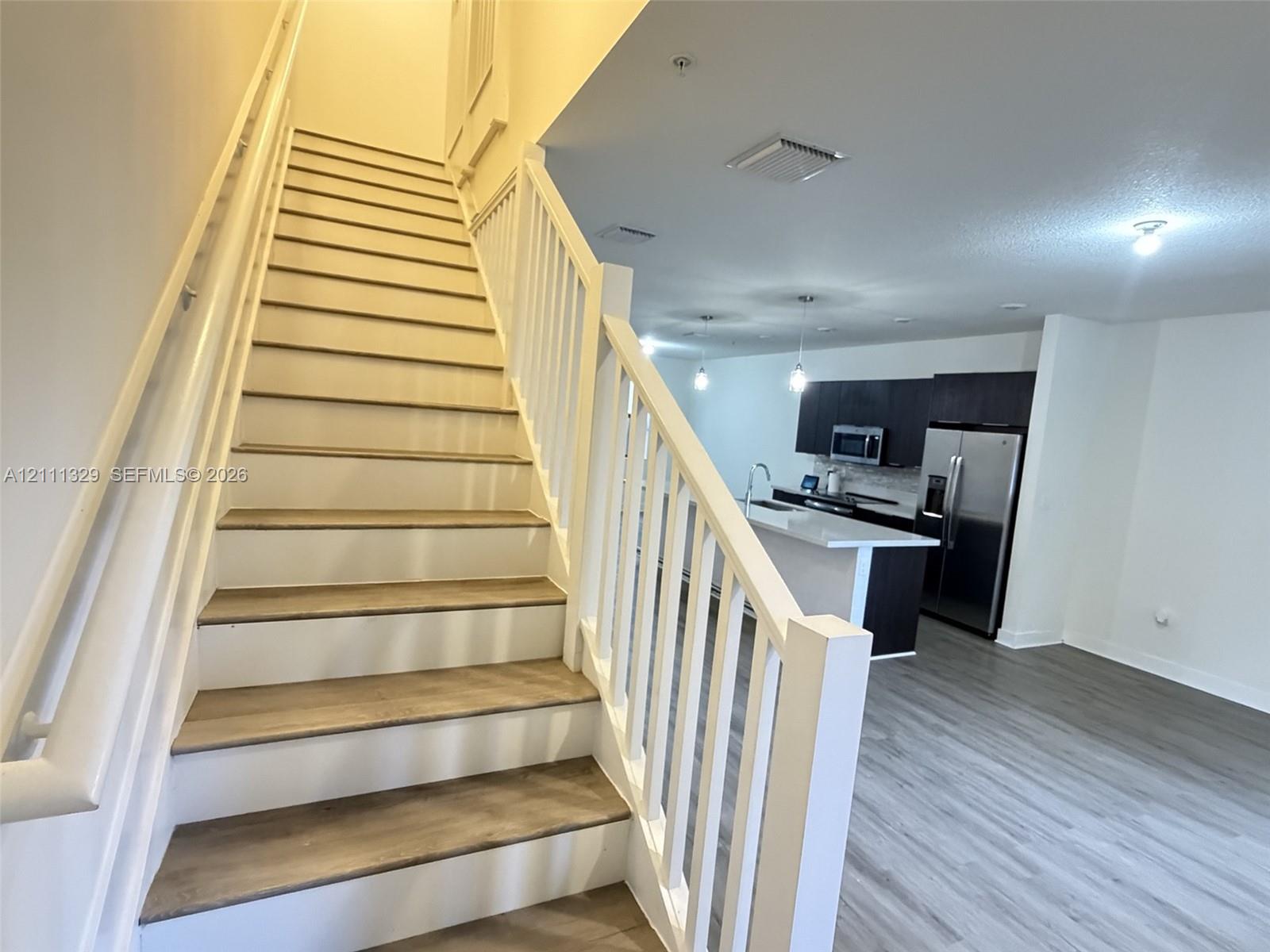 Staircase with wood-finish treads and white balusters, leading to an open-concept kitchen featuring dark cabinetry, an island with a light-toned countertop, and stainless steel appliances