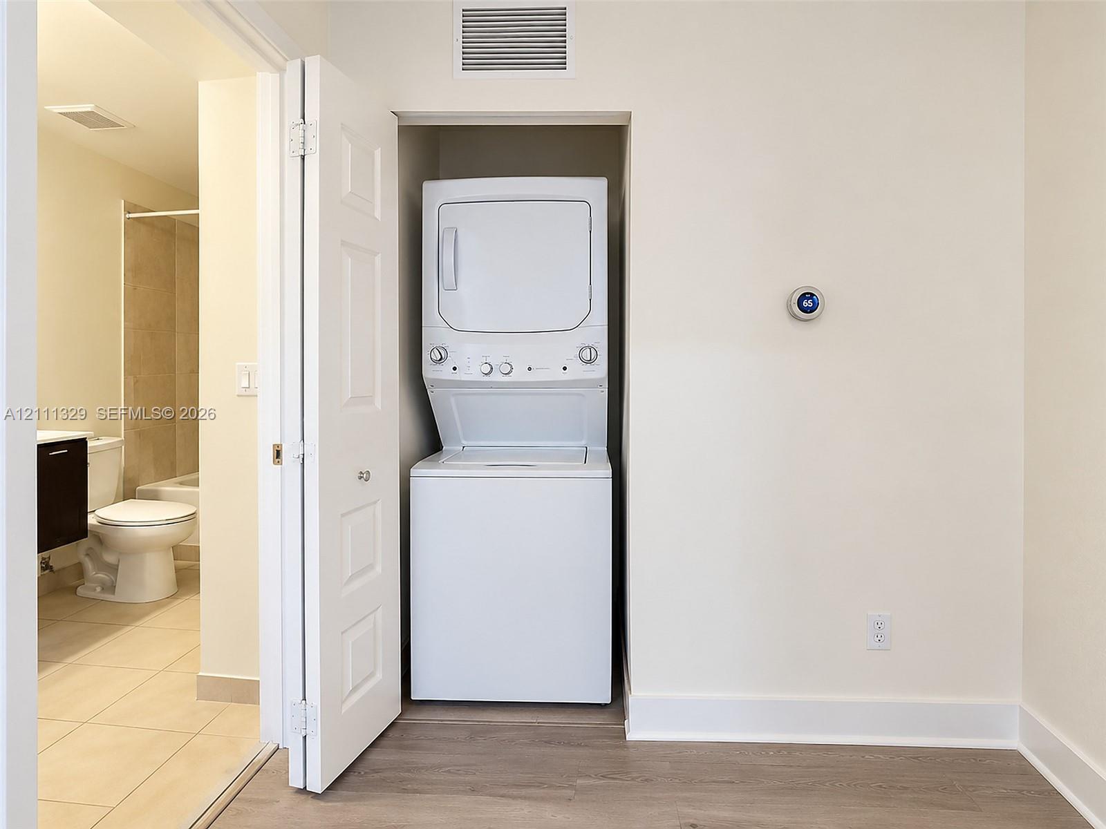 Laundry closet featuring a stackable washer and dryer unit, white six-panel bifold door, digital thermostat, and wood-finish flooring