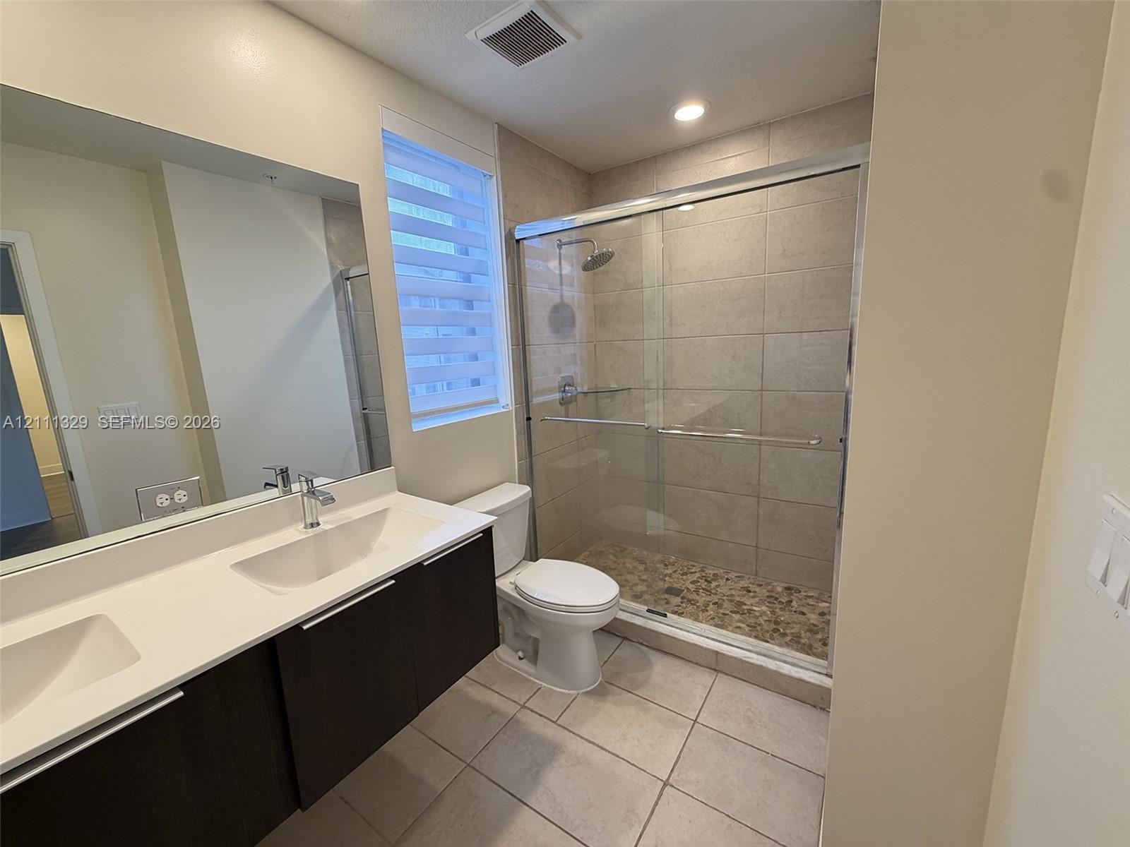 Double vanity featuring a light-toned countertop with integrated rectangular basins, dark wood-finish cabinetry, a large frameless mirror, and a walk-in shower with stone tile flooring and glass sliding doors