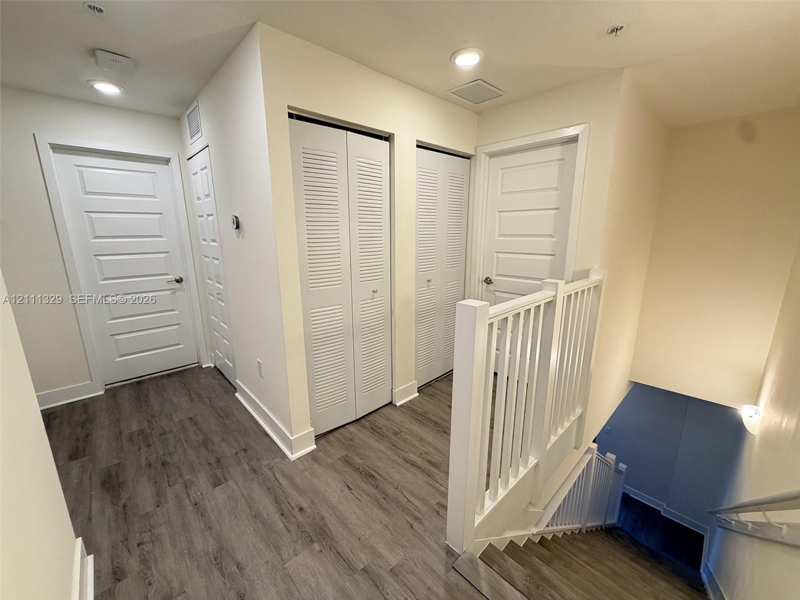 Hallway featuring wood-finish flooring, two sets of louvered-panel closet doors, recessed lighting, and a white balustrade staircase railing