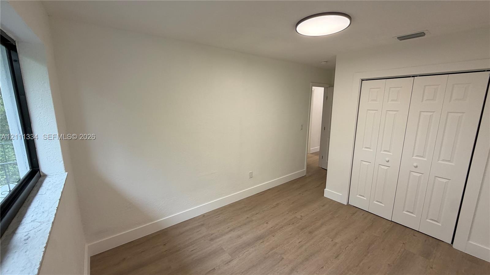 Interior space featuring wood-finish flooring, a large window with a deep sill, neutral wall finishes, a flush-mount circular lighting fixture, and a white bi-fold closet door
