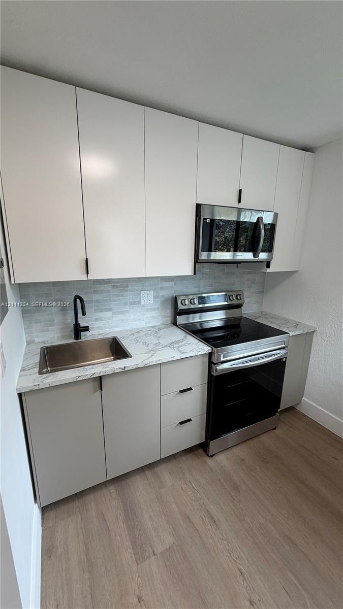Modern kitchen featuring white flat-panel upper cabinetry, light gray base cabinets, patterned stone-finish countertops, stainless steel appliances, and wood-finish flooring