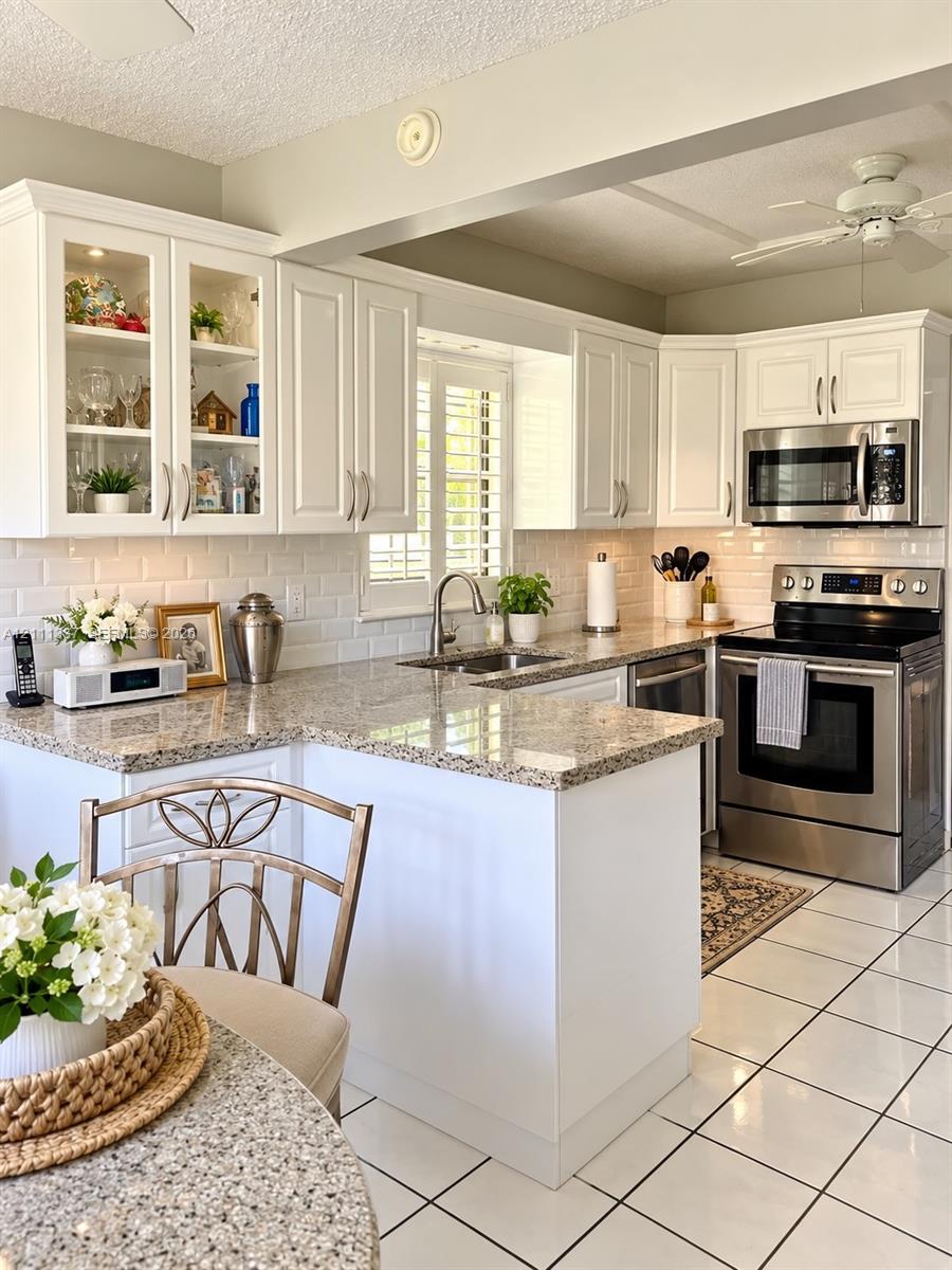White cabinetry with glass-front uppers, stone-finish countertops, subway tile backsplash, stainless steel appliances, and tiled flooring White cabinetry with glass-front uppers, stone-finish countertops, subway tile backsplash, stainless steel appliances, and tiled flooring