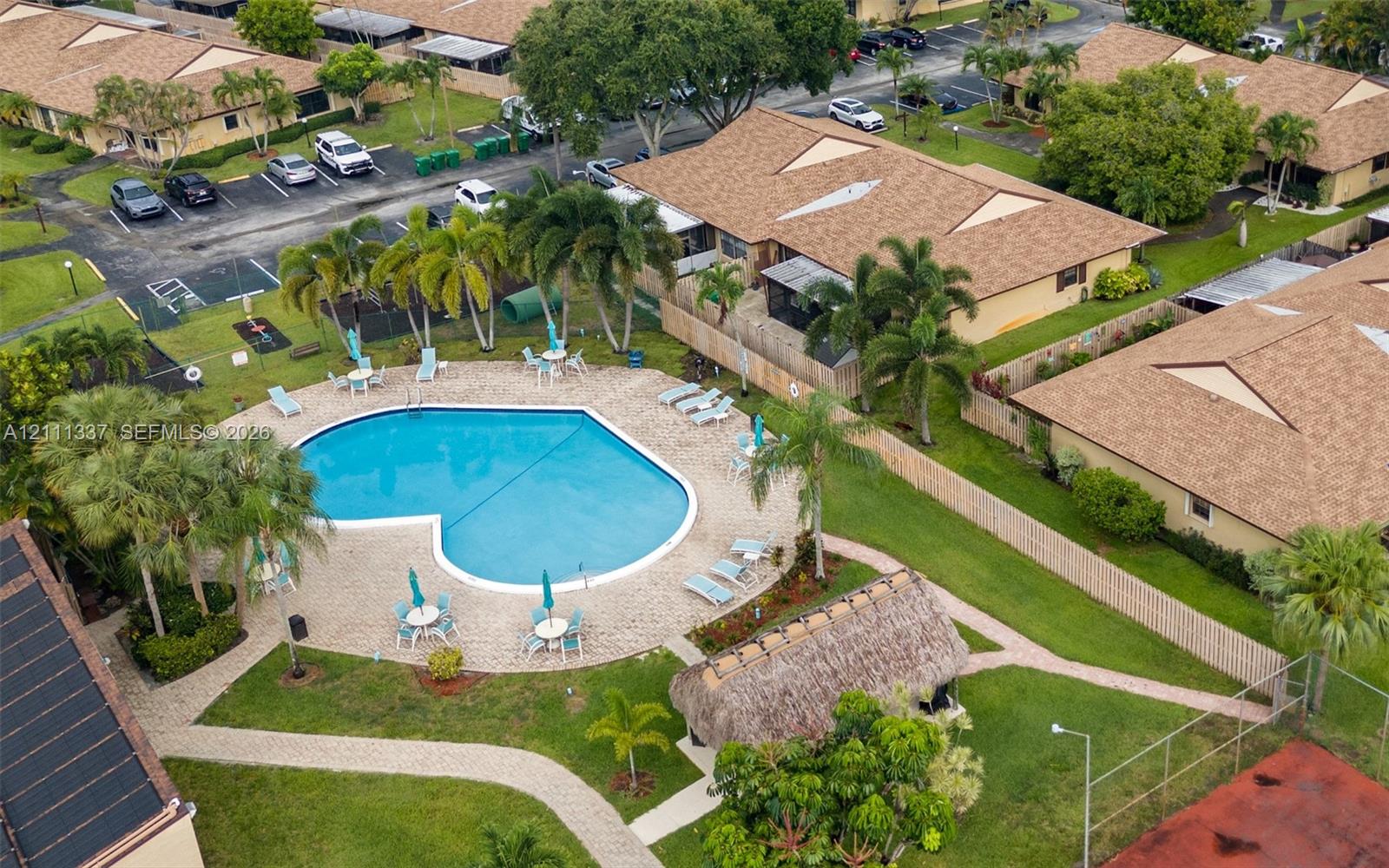 Community swimming pool featuring a paved deck, thatched-roof cabana, mature palm trees, and an enclosed tennis court Community swimming pool featuring a paved deck, thatched-roof cabana, mature palm trees, and an enclosed tennis court