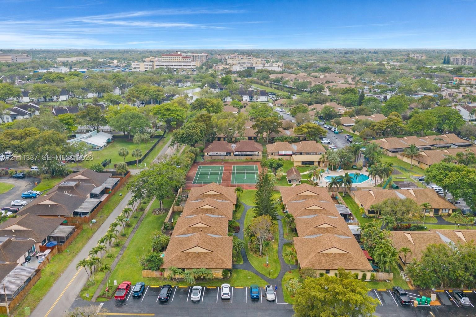 Aerial perspective of a multi-unit residential community featuring brown shingled roofing, dual tennis courts, a swimming pool, and dedicated paved parking areas