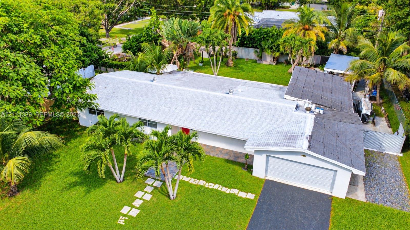 Single-story residence featuring a shingled roof, asphalt driveway, two-car garage, and expansive lot with mature tropical landscaping