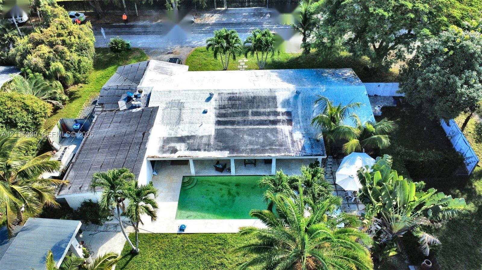 Aerial view of a residential property featuring an in-ground swimming pool, stone-paved patio, expansive lawn, and mature palm trees