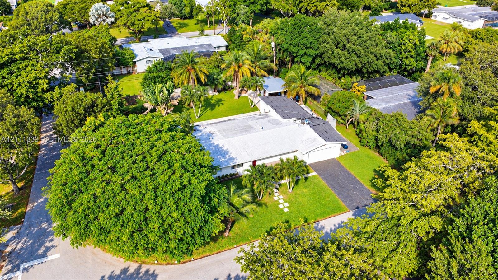 Aerial perspective of a single-story residence featuring a paved driveway, expansive lawn, mature canopy trees, and palm landscaping