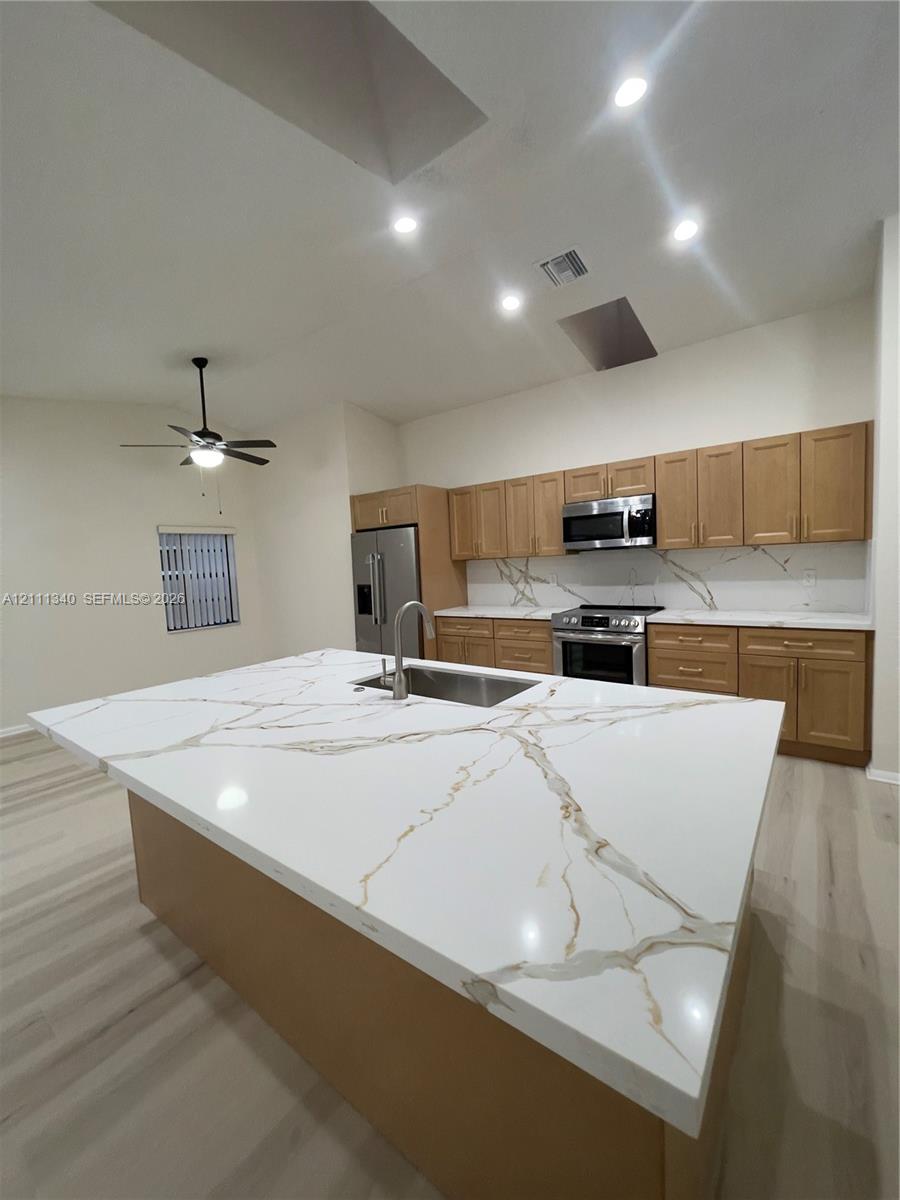 Kitchen featuring a large central island with veined stone-patterned countertops, wood-finish cabinetry, stainless steel appliances, recessed ceiling lighting, and wood-finish flooring Kitchen featuring a large central island with veined stone-patterned countertops, wood-finish cabinetry, stainless steel appliances, recessed ceiling lighting, and wood-finish flooring