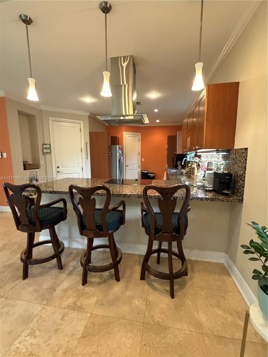 Kitchen featuring an expansive stone countertop, stainless steel ceiling-mounted vent hood, natural stone tile flooring, three pendant lights, and wood-finish cabinetry