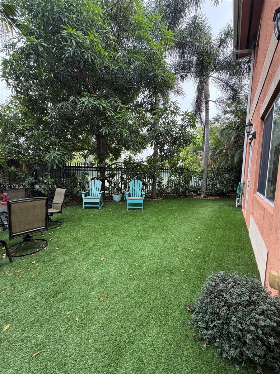 Private outdoor space featuring synthetic lawn, mature perimeter trees, established shrubbery, and metal fencing