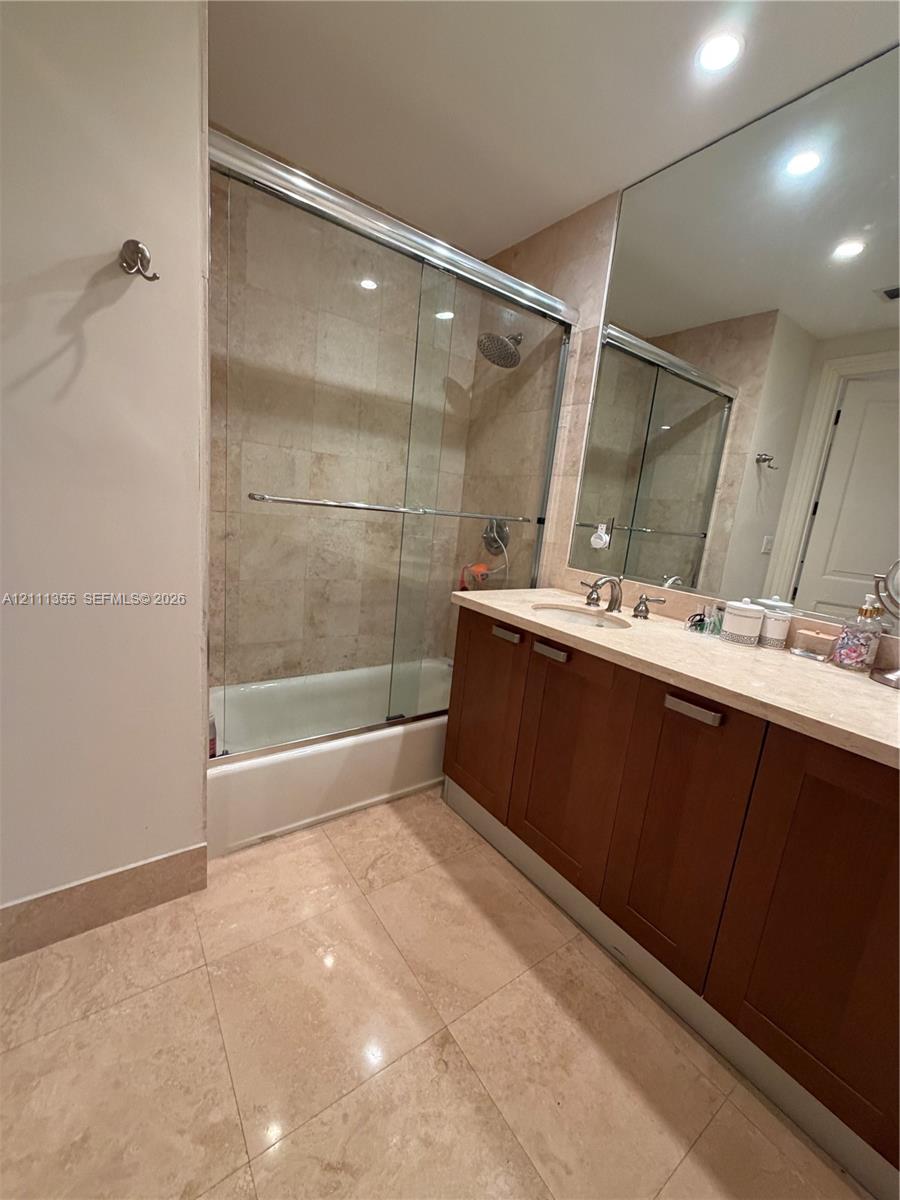 Bathroom featuring a vanity with wood-finish cabinetry, a light-toned stone-style countertop, a wall-spanning mirror, and a tub-shower combination enclosed by a glass sliding door with tiled wall surrounds