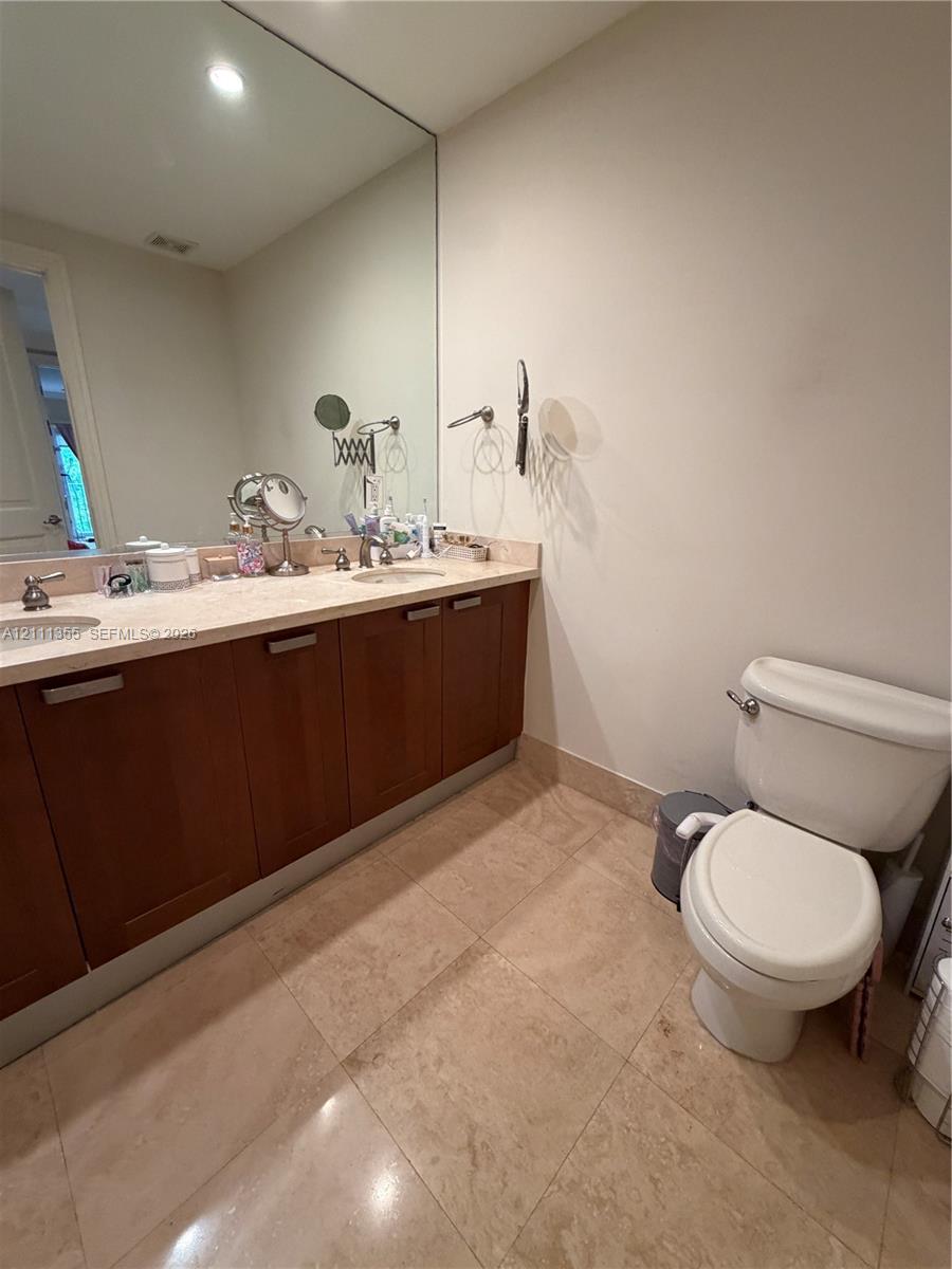 Dual-sink vanity with wood-finish cabinetry, solid surface countertop, expansive wall-to-wall mirror, and neutral tile flooring