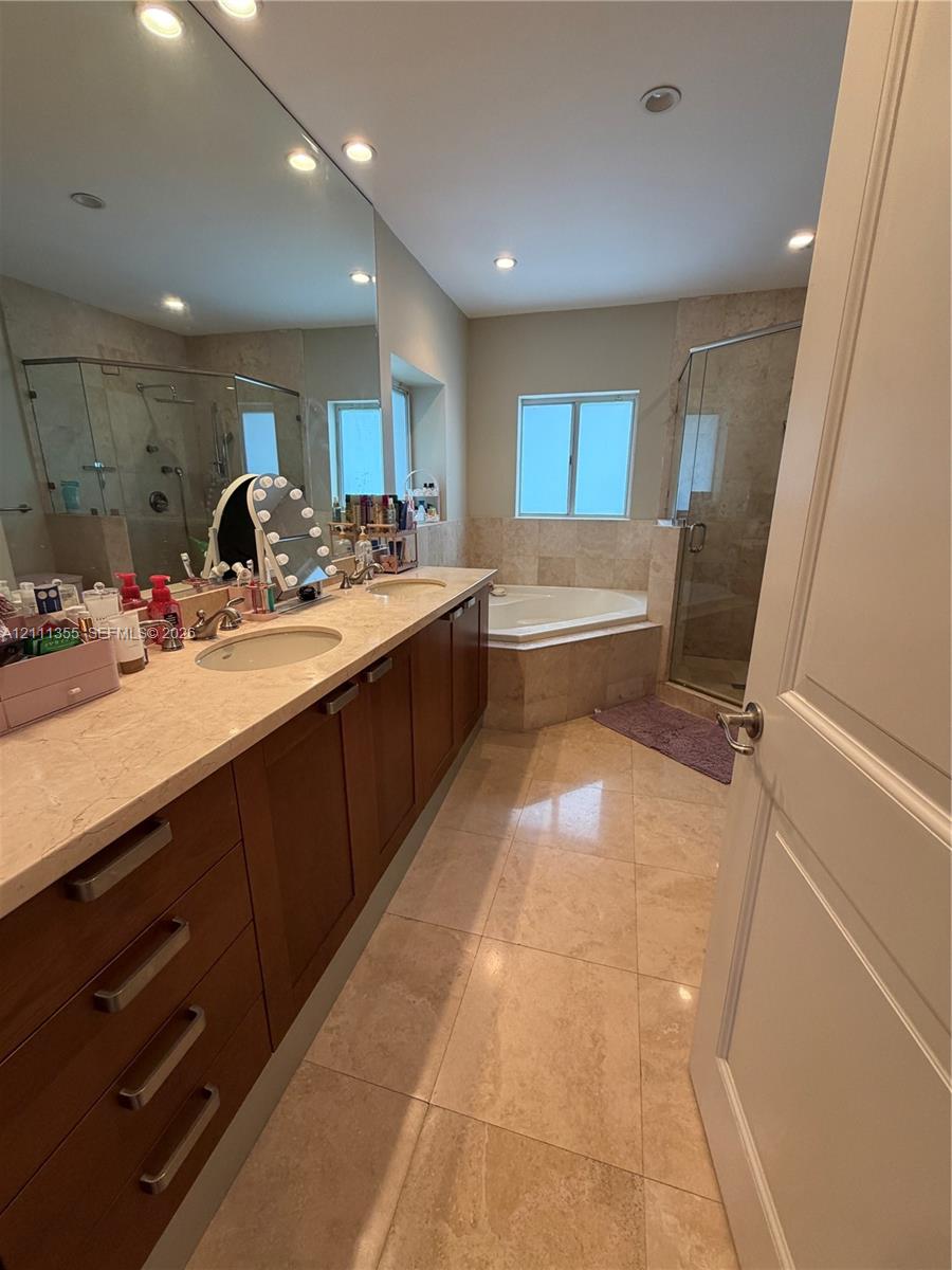 Dual-sink vanity with natural stone countertop, recessed lighting, full-width mirror, soaking tub with stone surround, and glass-enclosed shower