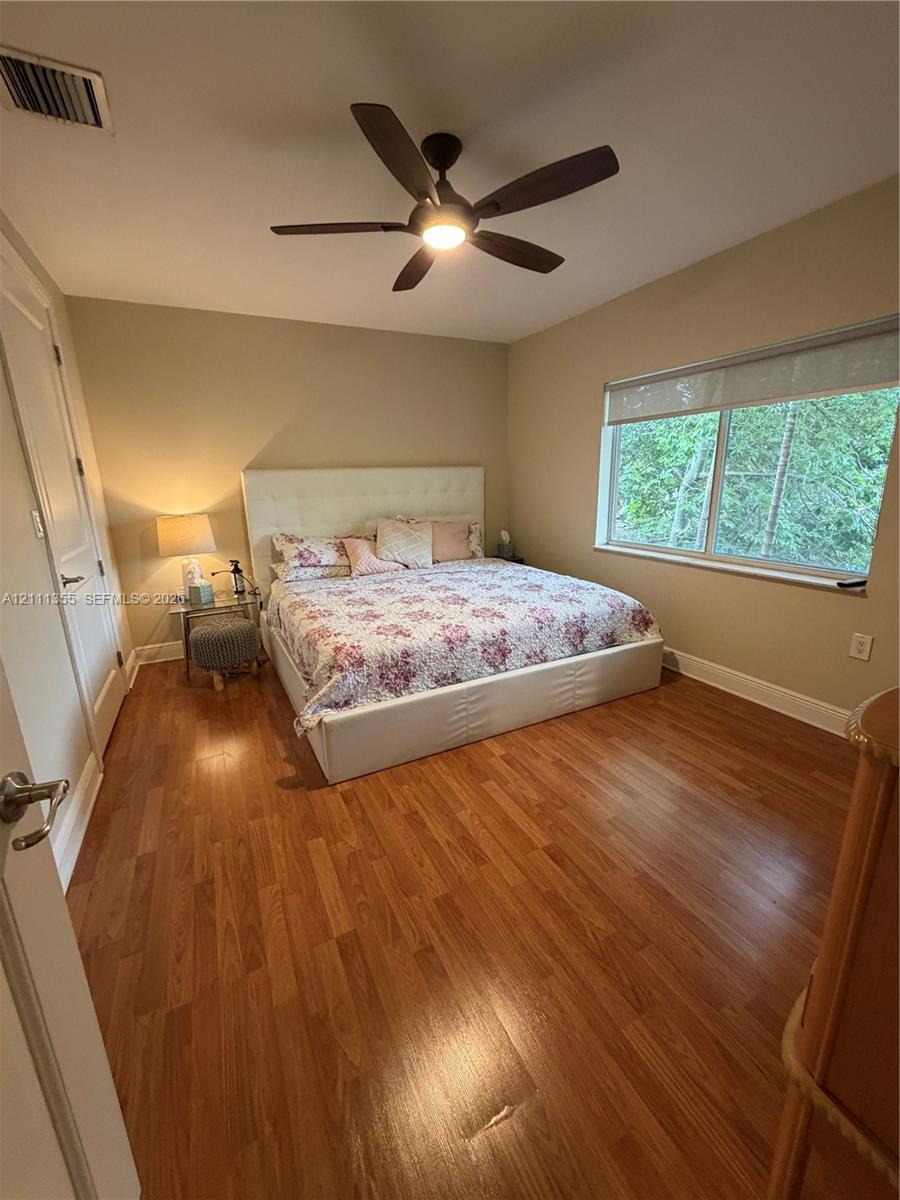 Room featuring wood-finish flooring, a multi-blade ceiling fan with integrated lighting, and a large window with a roller shade
