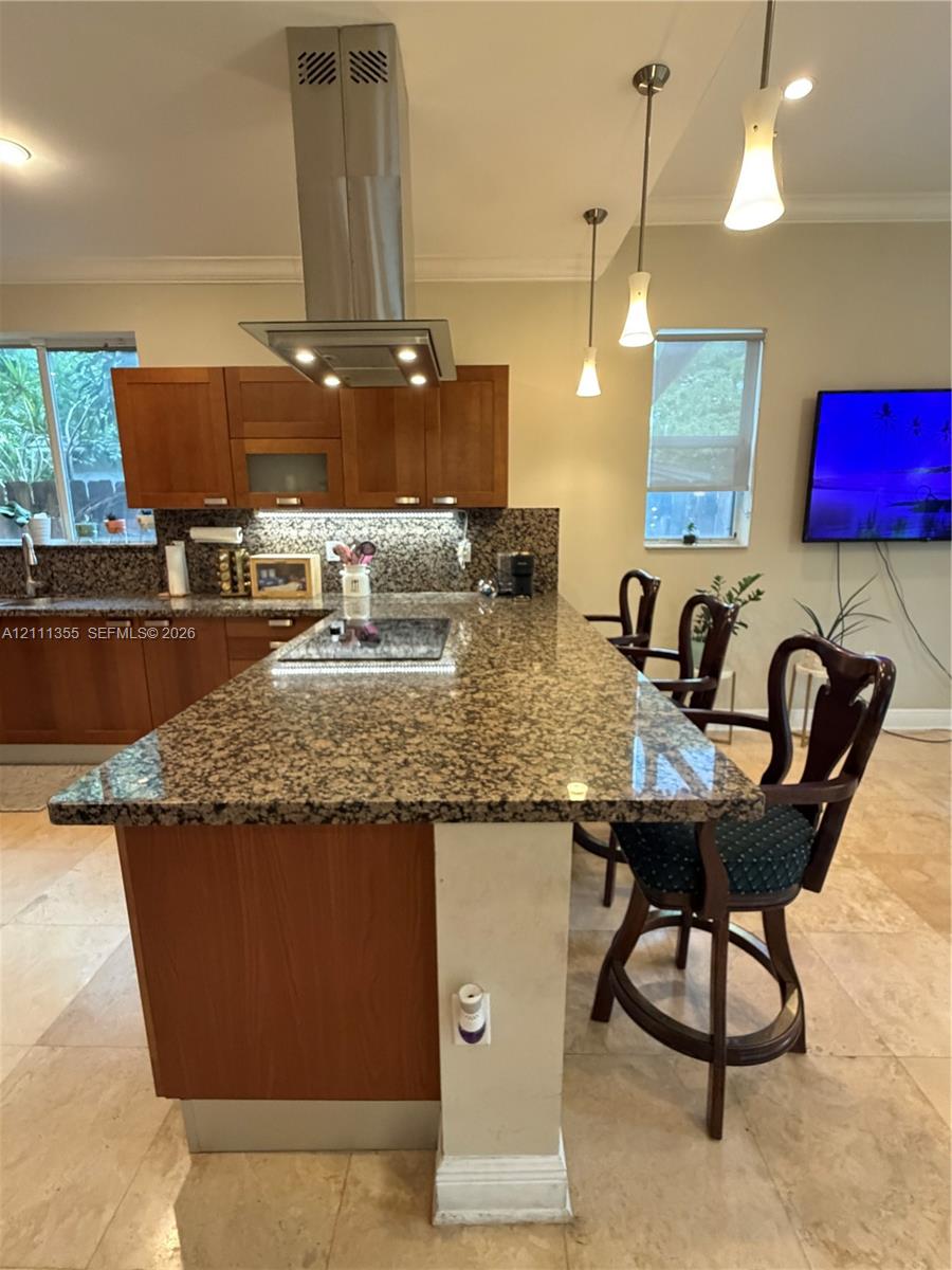 Kitchen featuring a sprawling stone-topped island with a built-in cooktop, stainless steel chimney-style range hood, warm-toned wood cabinetry, and pendant lighting