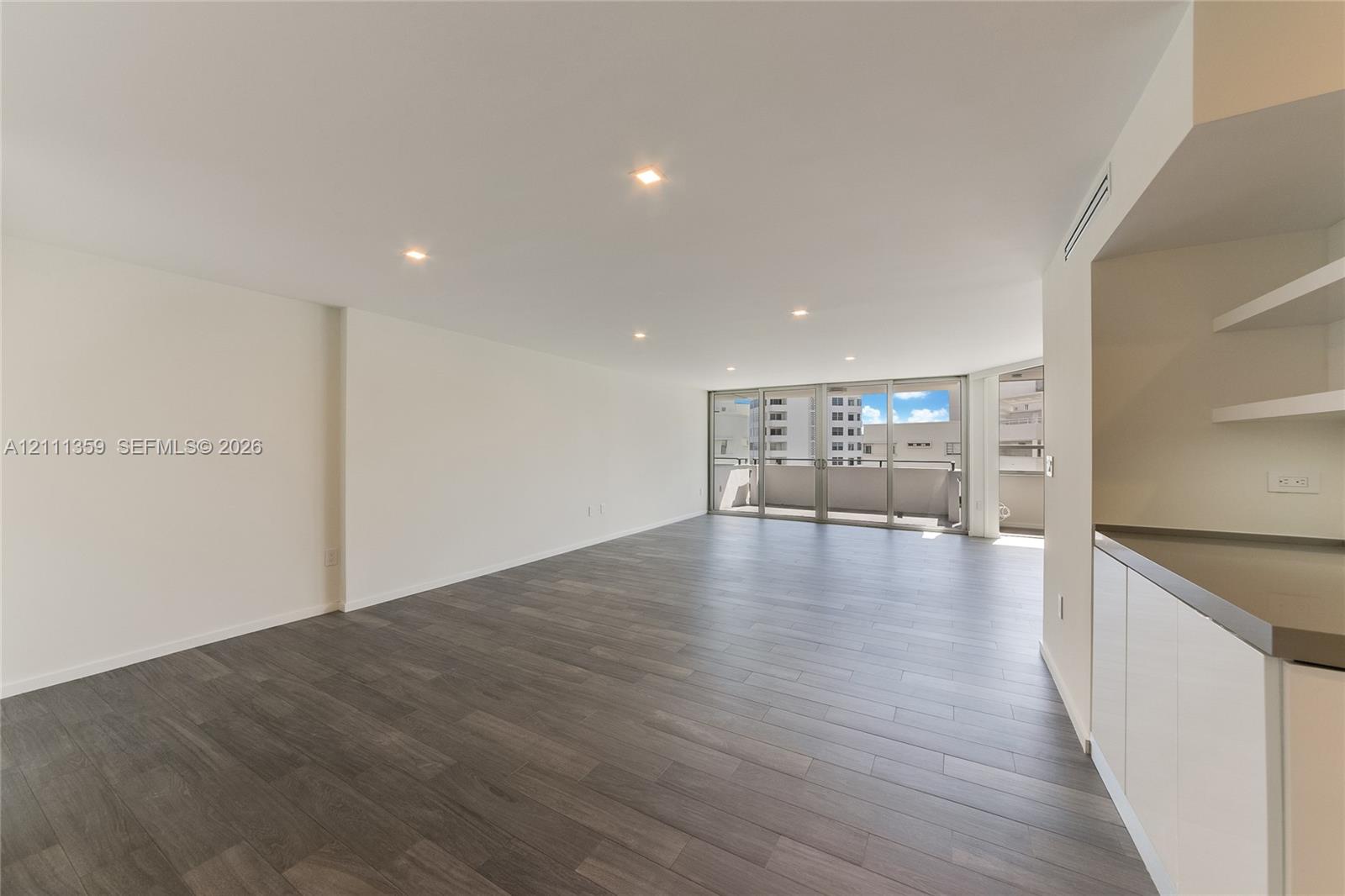 Open-concept interior featuring wood-finish flooring, recessed lighting, floor-to-ceiling glass sliding doors, private balcony, and built-in cabinetry with stone-finish countertops