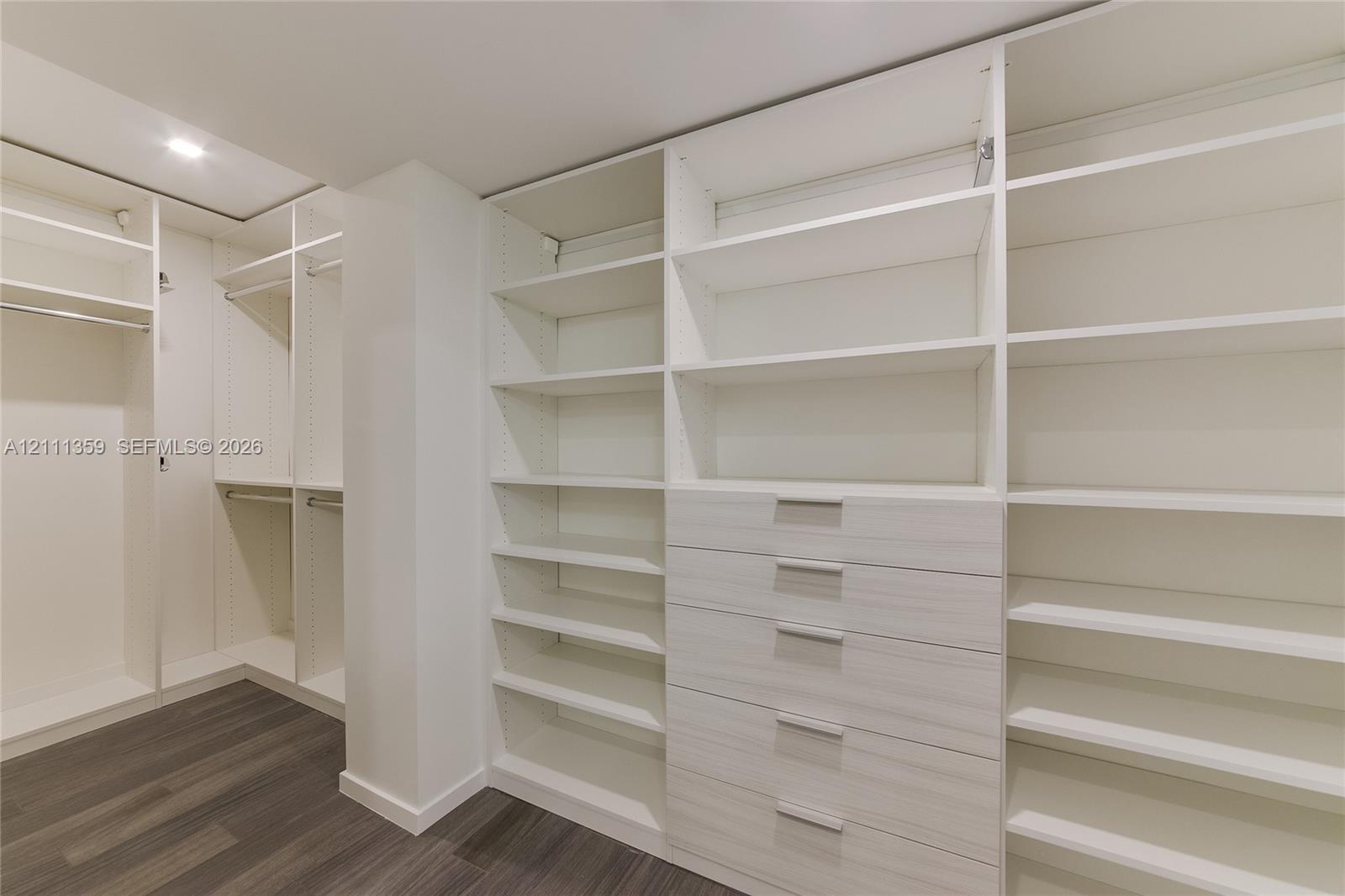 Walk-in closet featuring extensive built-in shelving, integrated wood-finish cabinetry, a bank of five drawers with metallic hardware, and dark wood-finish flooring