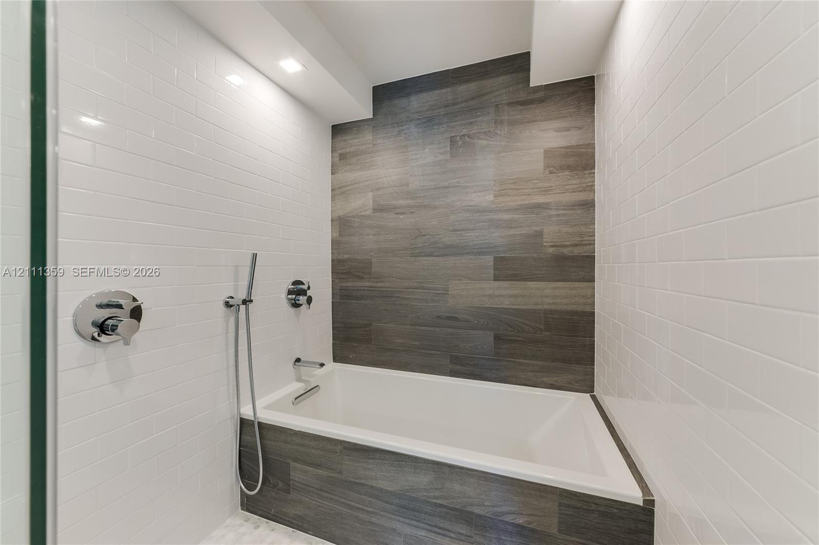 Built-in deep soaking tub featuring wood-finish tile surround, white subway tile walls, chrome shower fixtures, and recessed ceiling lighting